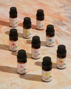 Nine small amber glass essential oil bottles with black caps and white labels are arranged on a warm beige marble-like surface. Each label features a different flower illustration in pink, yellow, blue, and orange, with the brand name 'FRANZIN' visible on the front.