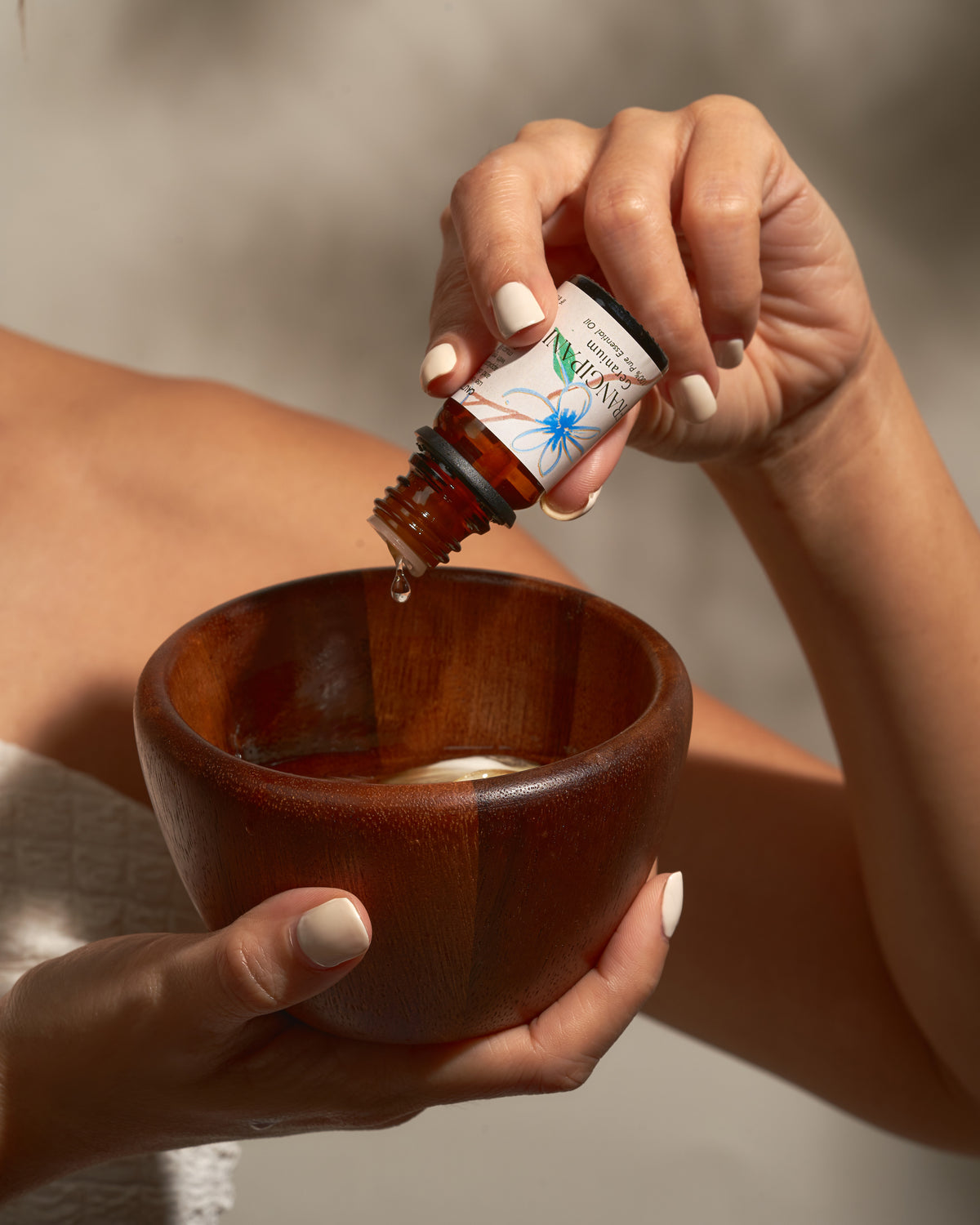A close-up of a person's hands holding a small amber glass bottle and pouring liquid into a wooden bowl. The bottle label reads 'Solei' and 'Rosemary & Rosehip'.