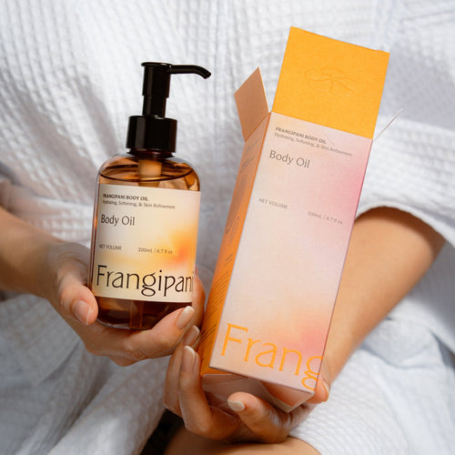 Person in a white robe holding Frangipani body oil beside its box