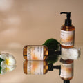 Two Frangipani body oil bottles with plumeria and moss on a reflective surface